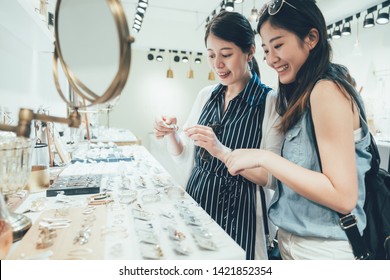 Two Asian Women Customer In Busy Clothes Shop Shopping Accessories Taking Up From Table. Beautiful Smiling Young Ladies Best Friends Buying Choosing Earrings On Desk In Fashion Modern Store Tokyo