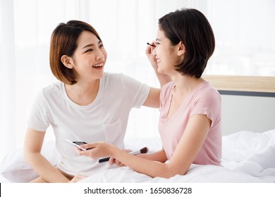 Two Asian women doing makeup for each other on bed in bright bedroom. Concept  for teenage or friends activity, lifestyle at home.