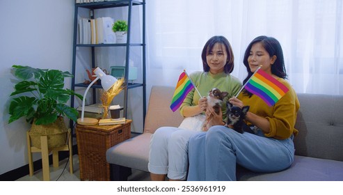 Two Asian women lesbian share a comforting tender moment smiles and a loving embrace on a sofa draped in a rainbow flag with two small dogs beside them in a cozy home - Powered by Shutterstock