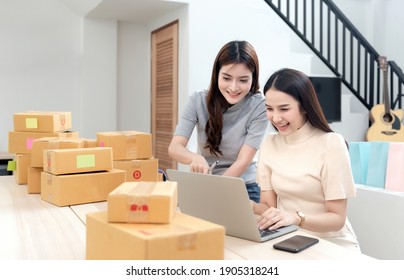 Two Asian Women Are Looking At Their Laptop Computers To Check Orders, With Cardboard Boxes And Shopping Bags All Around, Selling Products Online From Home. Being A New Normal Online Business