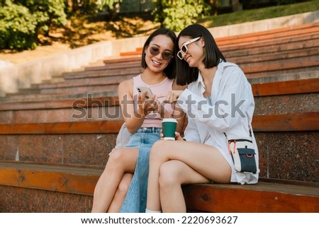 Two asian women smiling and using cellphone while sitting on bench outdoors