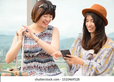 Two Asian Women Using App On Mobile Phone, Looking At Screen Of Smart Phone, Texting, Watching Photos While Traveling.