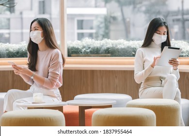 Two Asian women wearing mask to protect virus and keeping social distancing while using mobile phone and tablet in coffee shop . New Normal Concept. - Powered by Shutterstock