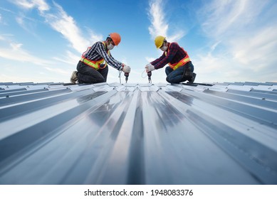Two Asian workers, roofing crews, wearing protective uniforms and gloves, using air or pneumatic nail guns and installing asphalt shingles on top of the new roof.