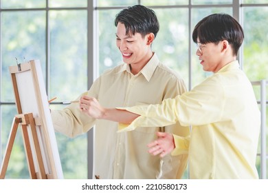 Two Asian Young Lovely Pride Teenager Male Gay Men Lover Couple Partner Standing Smiling Having Fun Playing Paintbrush In Front Canvas Painting Board On Easel In Glass Windows Workshop Studio At Home.