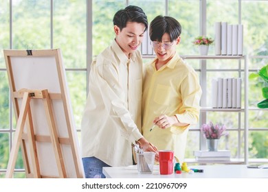 Two Asian Young Lovely Pride Teenager Male Gay Men Lover Couple Partner Standing Smiling Having Fun Playing Paintbrush In Front Canvas Painting Board On Easel In Glass Windows Workshop Studio At Home.