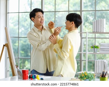 Two Asian Young Lovely Pride Teenager Male Gay Men Lover Couple Partner Standing Smiling Having Fun Playing Paintbrush In Front Canvas Painting Board On Easel In Glass Windows Workshop Studio At Home.