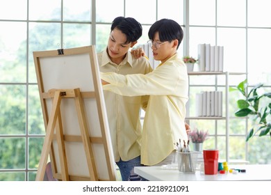 Two Asian Young Lovely Pride Teenager Male Gay Men Lover Couple Partner Standing Smiling Having Fun Playing Paintbrush In Front Canvas Painting Board On Easel In Glass Windows Workshop Studio At Home.