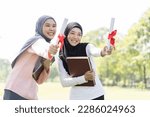 Two Asian young muslim woman wear hijab celebrating graduation ,holding book and degree scroll with red ribbon with happiness feeling, smiling in park. Education concept