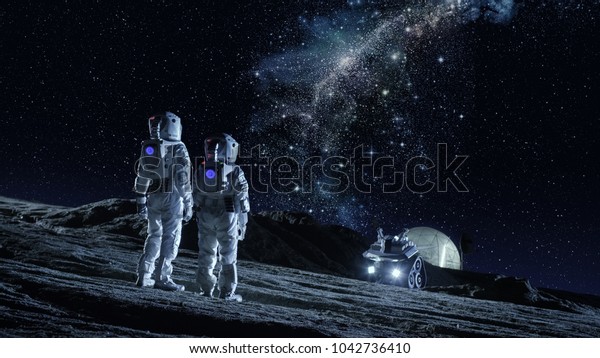 Two\
Astronauts in Space Suits Stand on the Planet and Looking at the\
The Milky Way Galaxy. In the Background Lunar Base with Geodesic\
Dome. Moon Colonization and Space Travel\
Concept.