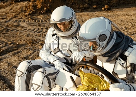 Two astronauts in space suits work together on a small vehicle while testing equipment on a terrain resembling Mars. The scene suggests a future exploration mission beyond Earth.