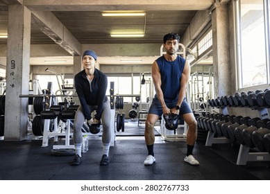 Two athletes in performance gear execute kettlebell squat inside modern gym with dumbbells, racks, and benches. Demonstrating proper form and strength training discipline, the movement highlights functional fitness, balance, and resilience. The scene captures realism of workout culture, emphasizing technique, training intensity, and structured exercise programming.