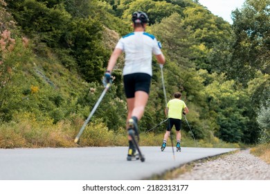 Two athletic men training on the roller ski at country road, back view. Defocused sportsman at foreground. Low angle view. Concept of sports competition and biathlon workout.