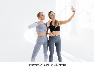 Two Athletic Sports Women Meet At Gym, Making Selfie Photo While Standing Together In The White Hallway. Meeting Friends And Having Fun At Gym