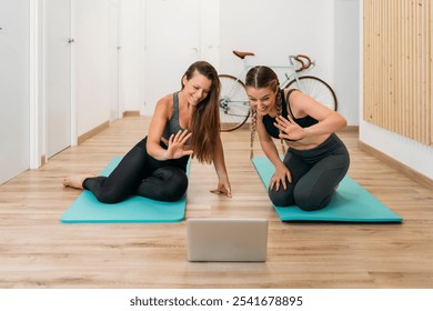 Two athletic women sitting on yoga mats are waving at a laptop during an online fitness class - Powered by Shutterstock