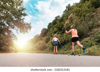 Two athletic women together training on the roller ski. Back low angle view. Copy space. Sunlight at finish. Concept of competition, biathlon, and summer workout.