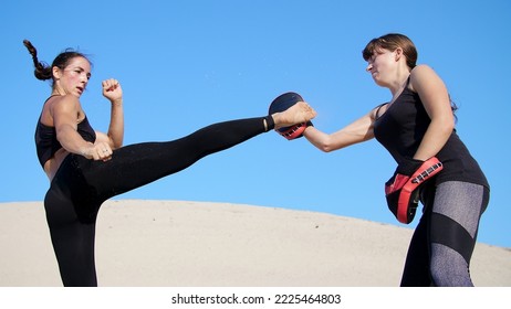 Two athletic, young women in black fitness suits are engaged in a pair, work out kicks, train to fight, on deserted beach, against a blue sky, in summer, under a hot sun. Slow motion. High quality - Powered by Shutterstock