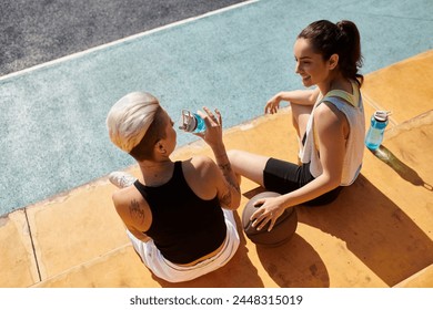 Two athletic young women sitting on the ground, hydrating with water bottles after playing basketball outdoors in the summer. - Powered by Shutterstock