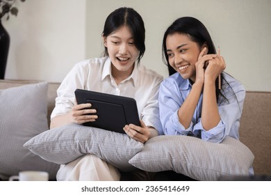 Two attractive Asian women using digital tablet with friends sitting on sofa at home. - Shutterstock ID 2365414649