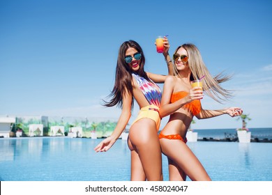 Two attractive brunette and blonde girls with long hair are posing near pool on the sun. They wear swimsuit, sunglasses and holding cocktails. They are moving.