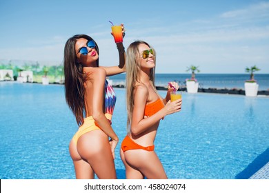 Two attractive brunette and blonde girls with long hair are posing near pool on the sun. They wear swimsuit with sunglasses. They have chilling time.
