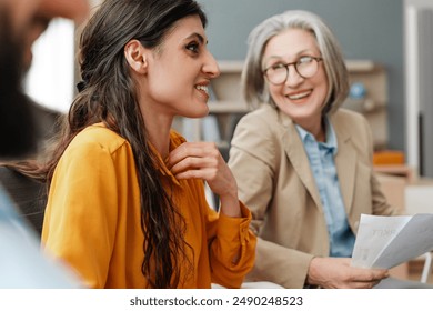 Two attractive businesswomen smiling and having conversation during meeting with colleagues, one of them is holding some paperwork. Communication concept - Powered by Shutterstock