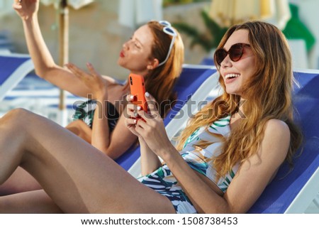 Two attractive fit girls wearing swimsuits laying on beach lounger at the swimming pool, holding mobile phones, takind selfies