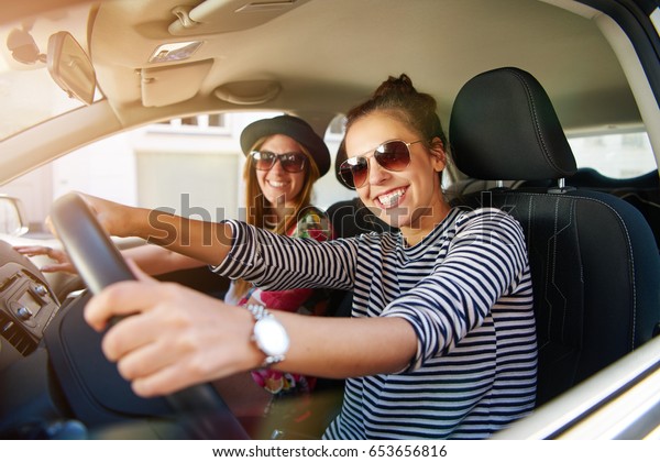Two attractive friendly young women enjoying a day\
trip to town viewed through the open window of their car grinning\
happily at the camera