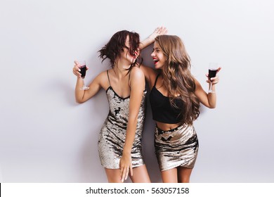 Two attractive girls, cheerful best friends dancing, having fun and drinking red wine on party. Wearing shiny dresses with paillettes, fashionable looking with beautiful wavy hair. Isolated.