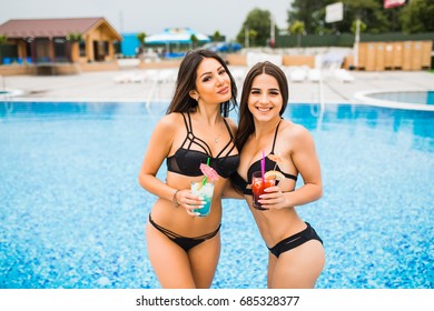 Two attractive girls with long hair are posing near pool on the sun. They wear swimsuit. They have chilling time.