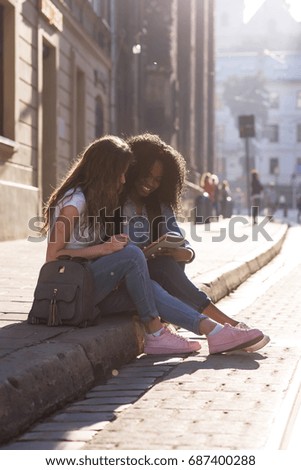 Two attractive girls sitting on the pavement and looking at the screen of the phone. one girl is african american. Beautiful city around.