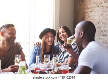 Two Attractive Ladies In Hat And Striped Shirt Laughing While Embarrassed Male Friends Drink Wine In Restaurant With Large Bright Window