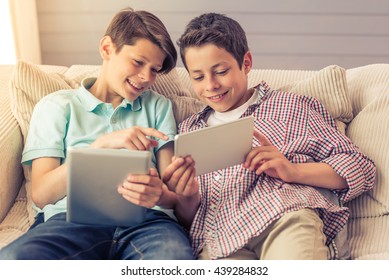 Two Attractive Teenage Boys Are Using Tablets, Talking And Smiling While Sitting On The Couch At Home