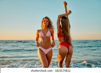 Two attractive women friends have fun on the beach in bikini swimwear at sunrise.