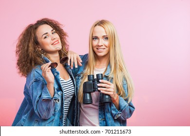 Two attractive women in jeans jackets with sunglasses and binoculars. Travel and vacation concept. Pink background studio shot.