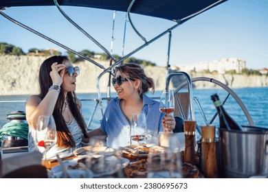 Two attractive women in sunglasses and casual clothes are having lunch on deck of ship. Handsome girls are laughing and drinking wine on boat. Pretty young ladies are having fun on sea voyage on yacht - Powered by Shutterstock