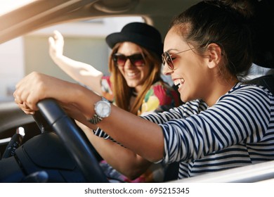 Two Attractive Young Friends Wearing Sunglasses Laughing Together While Driving In A Car Through The City On A Sunny Day