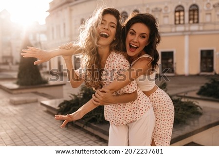 Two attractive young women fooling around in fresh air. Brunette and blonde with their mouths wide open are laughing, dressed in casual light clothes. Summer playful mood concept