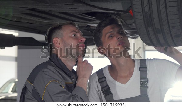 Two auto mechanics checking
suspension of a lifted car at the garage. Car service technicians
repairing automobile at their workshop. Service, experience
concept