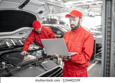 Two auto mechanics in red uniform doing engine diagnostics with computer in the car service - Powered by Shutterstock