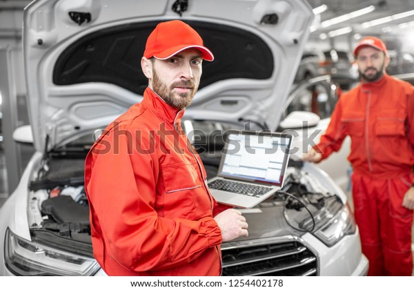 Two auto mechanics in red\
uniform doing engine diagnostics with computer in the car\
service
