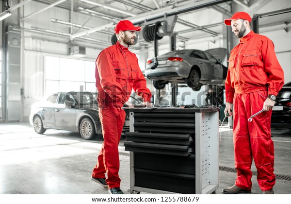 Two auto mechanics in red uniform walking with\
working cart at the car\
service