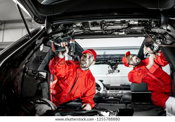 Two auto service workers\
in red uniform disassembling new car interior making some\
improvements indoors