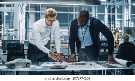 Two Automotive Engineers Working In Office At Car Factory. Industrial Designer Shows Conceptual Electric Engine Parts To Colleague, Discussing Different Applications In Technological Laboratory.