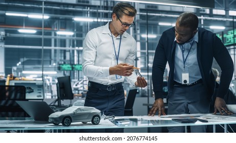 Two Automotive Engineers Working In Office At Car Factory. Industrial Designer Shows Conceptual Electric Engine Parts To Colleague, Discussing Different Applications In Technological Laboratory.