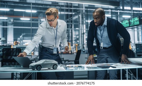 Two Automotive Engineers Working In Office At Car Factory. Industrial Designer Shows Conceptual Electric Engine Parts To Colleague, Discussing Different Applications In Technological Laboratory.