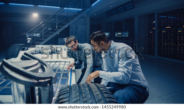 Two Automotive Engineers Working on Electric Car\
Chassis Platform, Taking Measures, working with 3D CAD Software,\
Analysing Efficiency. Vehicle Frame with Wheels, Engine and\
Battery.