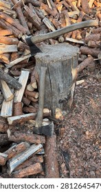 Two axes are stuck in a weathered chopping block, surrounded by a large pile of rough, split firewood logs. This rustic outdoor scene suggests winter preparation, manual labor, and sustainable home heating.
