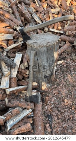 Two axes are stuck in a weathered chopping block, surrounded by a large pile of rough, split firewood logs. This rustic outdoor scene suggests winter preparation, manual labor, and sustainable home heating.