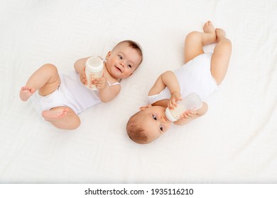 two babies suck milk from a bottle lying in a crib in the children's room on a white bed